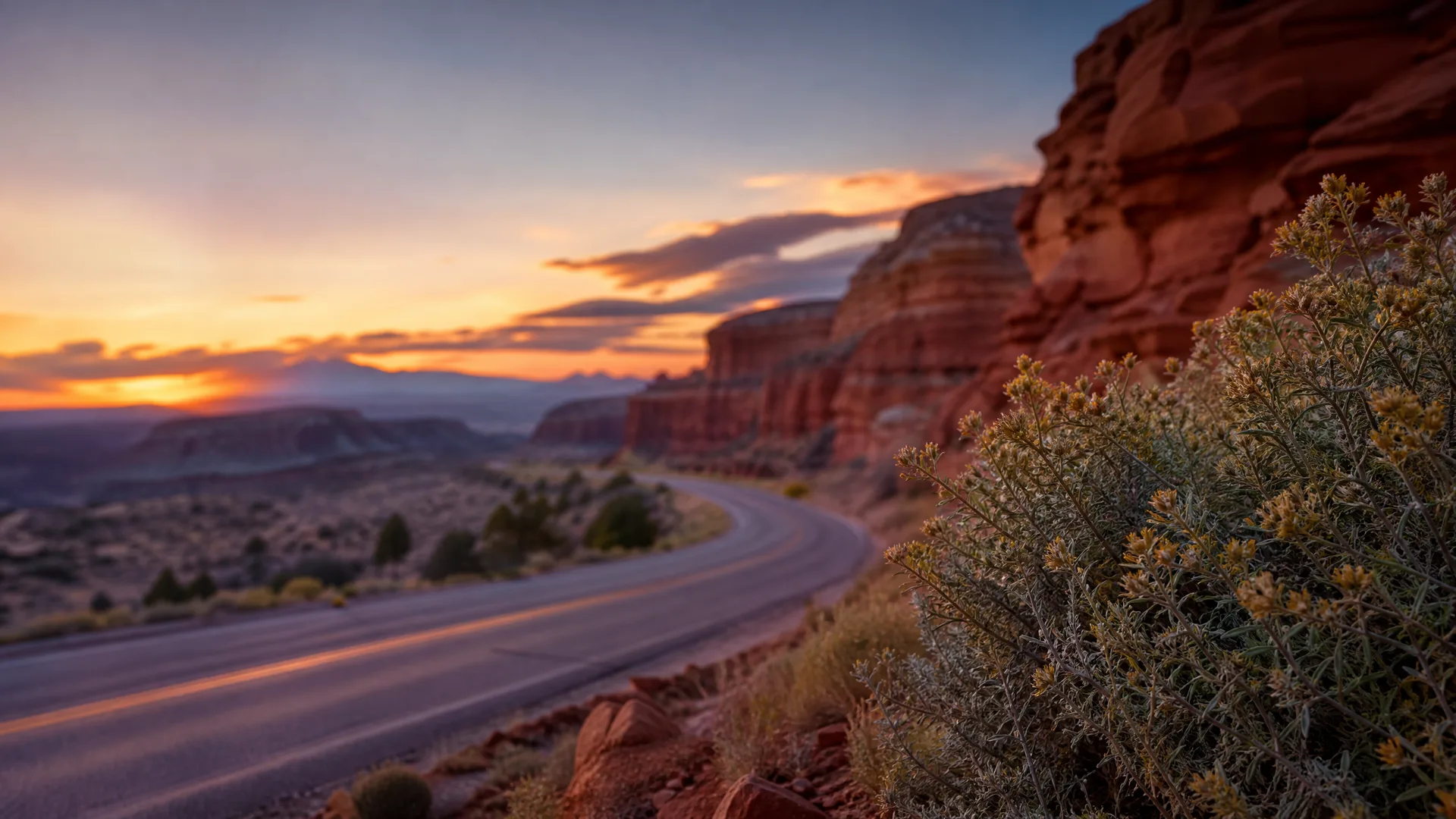 A two-lane road curves past red sandstone cliffs and desert scrub at sunset, with distant mesas and an orange-lit sky.