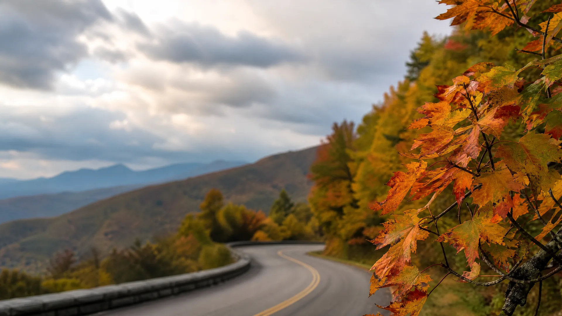 A curving two-lane road with a yellow center line and a low stone guardrail runs through forested hills, with orange and yellow maple leaves in sharp focus in the foreground and distant mountain ridges under a cloudy sky.