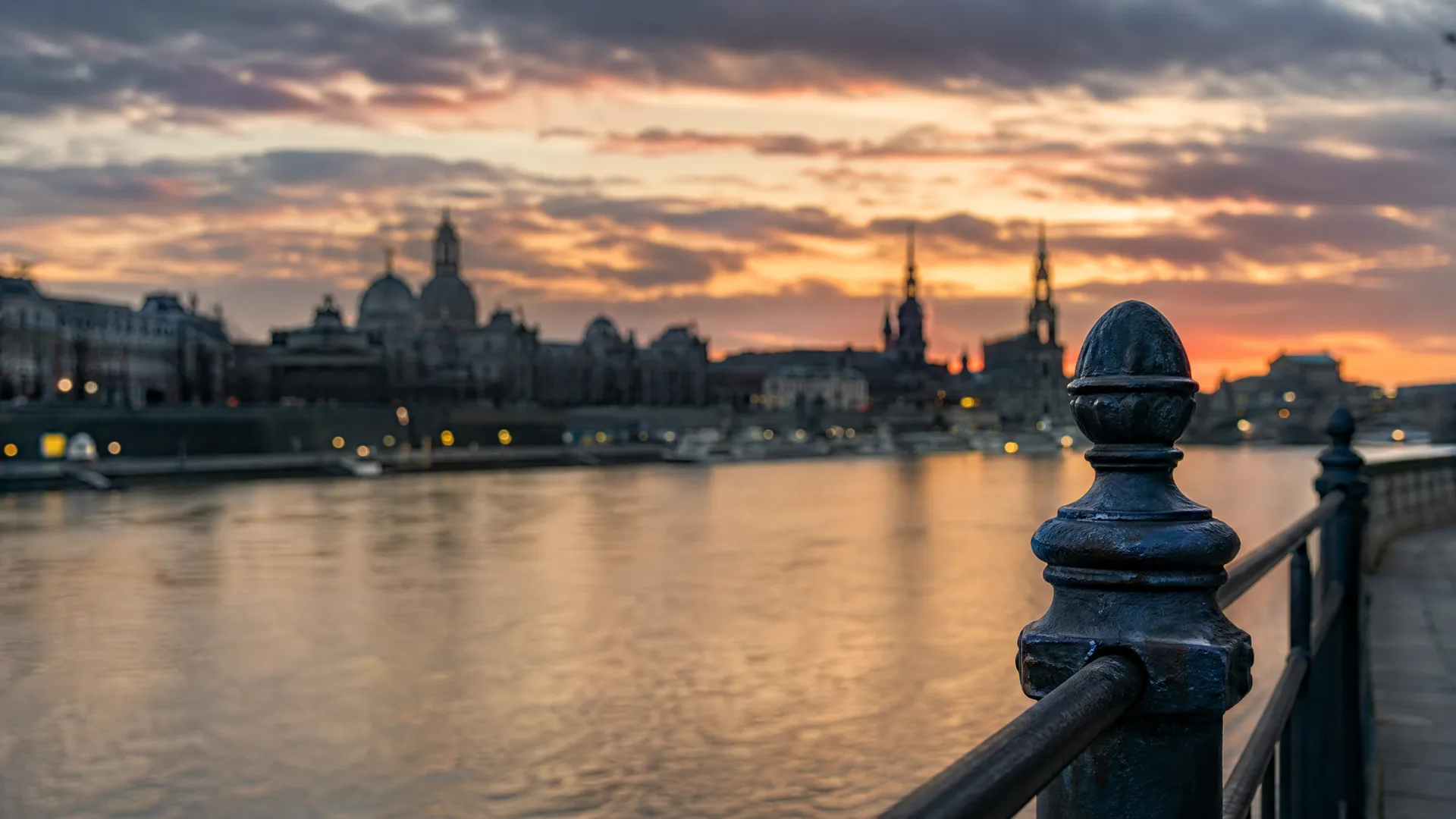 A dark metal railing post in sharp focus in the foreground, with a blurred river and city skyline of domes and towers under an orange and grey sunset sky behind.