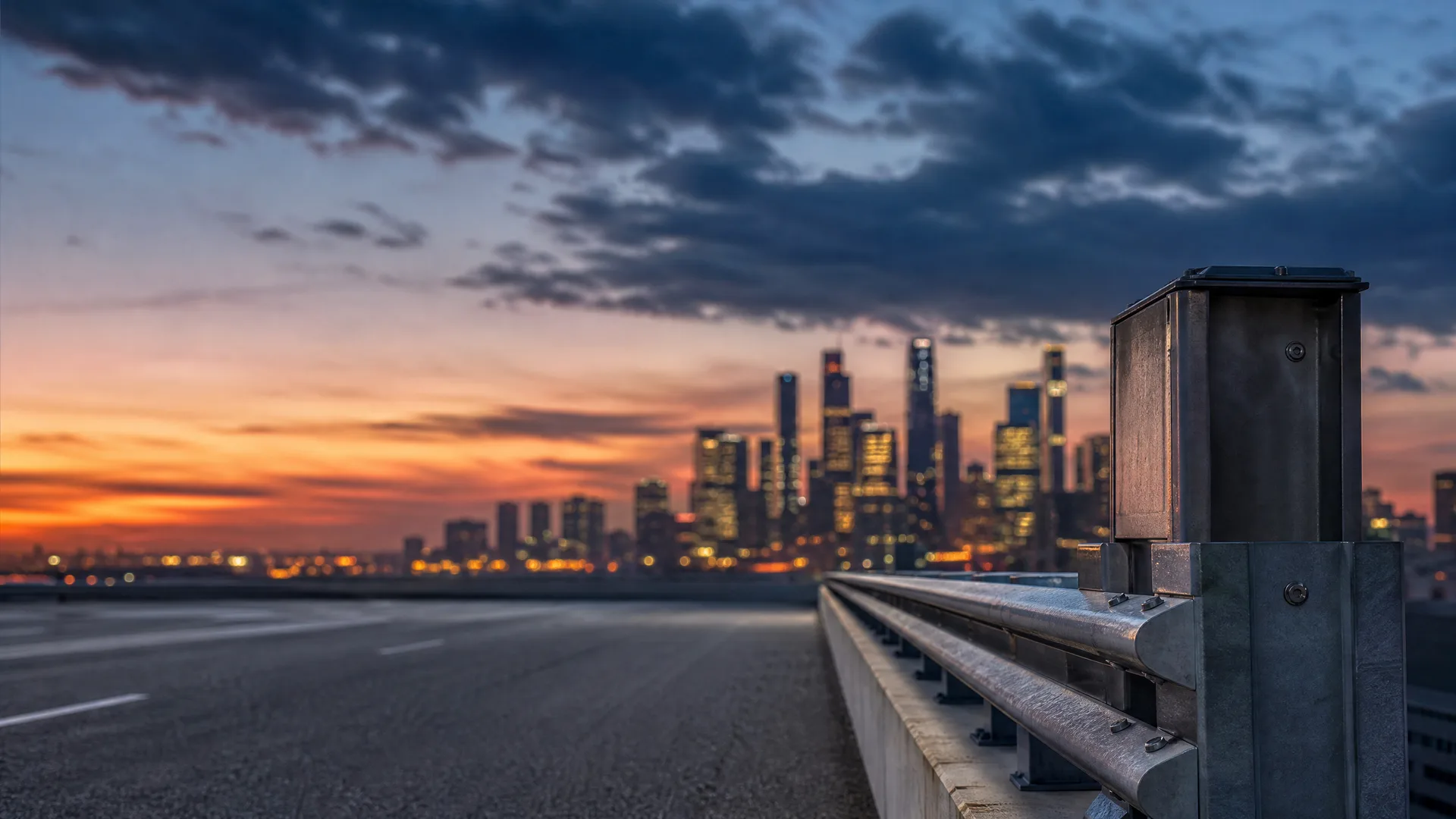 An empty asphalt road with a metal guardrail in the foreground and a blurred city skyline under an orange and blue sunset sky.