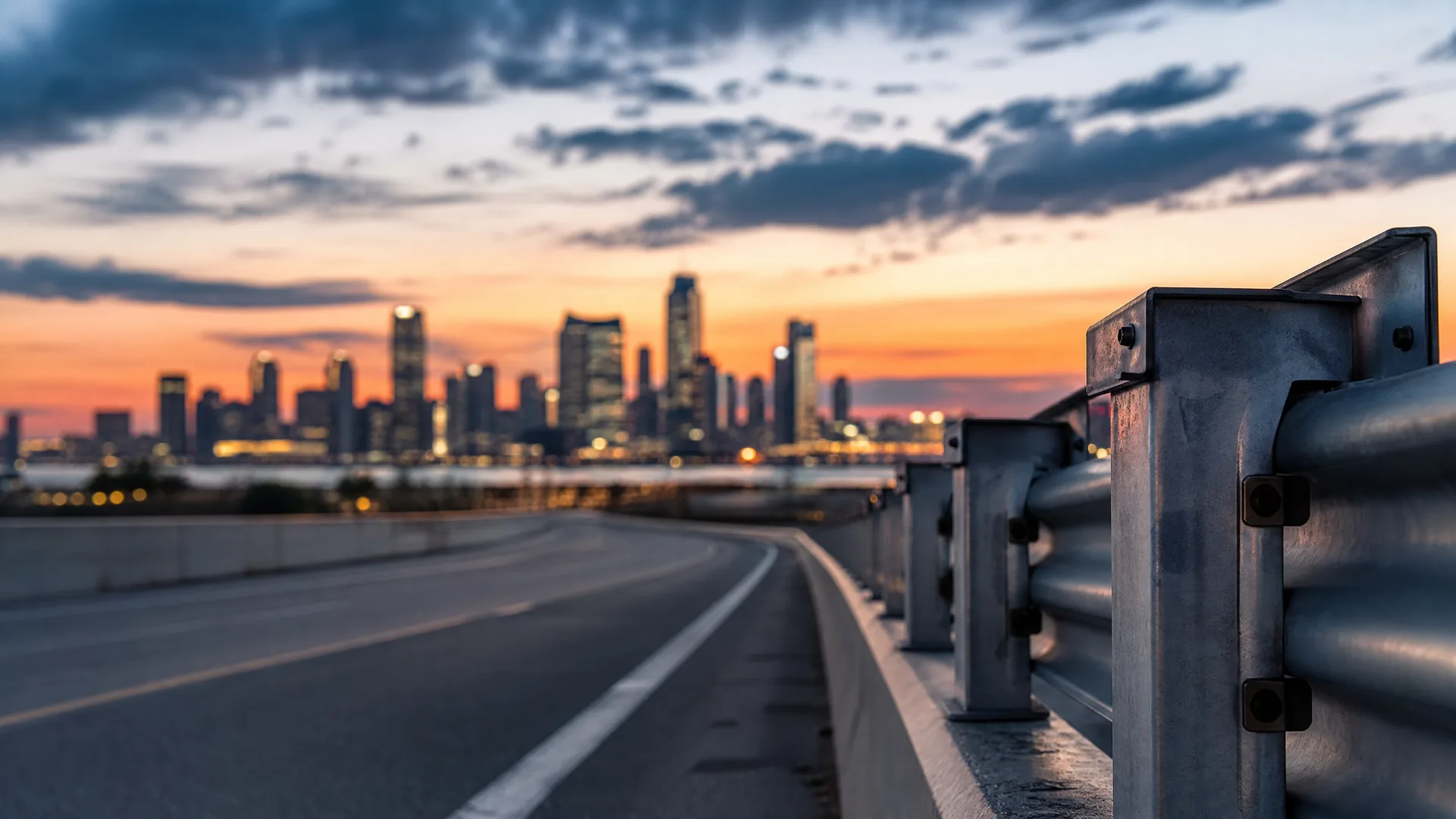 A curving asphalt road with a white lane line and a metal guardrail in sharp foreground focus, with a blurred city skyline under an orange and cloudy sunset sky in the background.